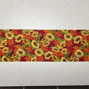 May include: A decorative table runner featuring a vibrant autumn floral pattern. The design includes sunflowers, red and orange leaves, and green foliage on a black background. The runner has a unique hexagonal shape.
