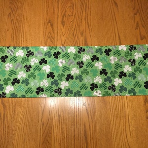 May include: Green table runner with a shamrock pattern. The shamrocks are in various shades of green, black, and white.