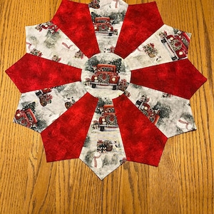 May include: A red and white Christmas table runner with a snowflake design and a red truck carrying a Christmas tree. The truck is surrounded by snowmen and other Christmas decorations.