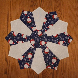 May include: A white and blue Christmas table runner with a snowflake design and a repeating pattern of Santa Claus figures. The runner is made of fabric and has a glittery finish.