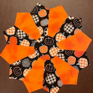 May include: A black and orange fabric table runner with a pumpkin pattern. The runner is made of multiple fabric pieces sewn together in a starburst pattern.