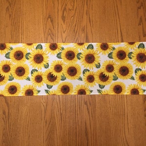 May include: A white table runner with a sunflower pattern. The runner is approximately 14 inches wide and 72 inches long.