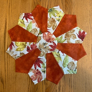 May include: A round, orange and white fabric placemat with a leaf pattern. The placemat is made of multiple fabric pieces sewn together in a starburst pattern.