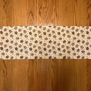 May include: A white table runner with a repeating pattern of brown and gray teddy bears. The runner has a pointed end on each side.