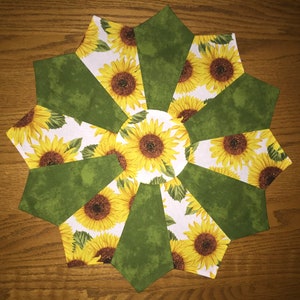 May include: A handmade, circular fabric item featuring a sunflower design. The center and alternating sections display vibrant yellow sunflowers on a white background. The other sections are a solid green color. The item is likely a decorative placemat or coaster.