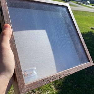 May include: A wooden shadow box with a glass front, displaying a white canvas backing. The frame is engraved with "Nicolle & Cade 5/21/2025". A small plastic bag containing jewelry is visible inside the box.