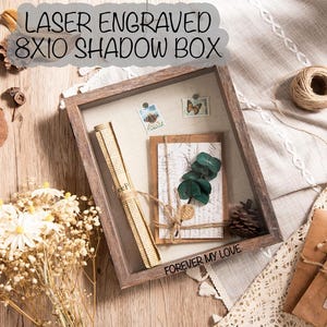 May include: A wooden shadow box with the text "LASER ENGRAVED 8X10 SHADOW BOX" and "FOREVER MY LOVE". Inside the box are decorative elements including a rolled paper, stamps, a card with greenery, and a pinecone. The box is 8 inches by 10 inches.