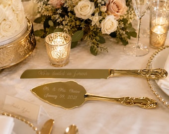 May include: A gold-toned cake knife and server set with ornate handles. The knife blade is engraved with the text "We've decided on forever," and the server is engraved with "Mr. & Mrs. Suarez" and the date "January 09, 2021" in cursive script.