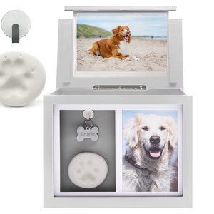 May include: White wooden box with a hinged lid, containing two photo frames and a paw print. The box has a small metal hook on the side. The photo frames are empty, but one has a dog tag with the name "Charlie" on it. The box is designed to hold pet mementos.