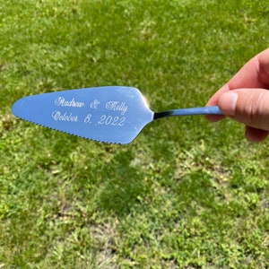 May include: Silver cake server with a serrated edge and a handle. The server is engraved with "Andrew & Kelly October 8, 2022".
