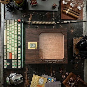 May include: A dark wood humidor with a glass top, a gold digital hygrometer, and the engraved text 'It's dangerous to go alone! Take this!' on the front. The humidor is surrounded by a mechanical keyboard, a computer mouse, cigars, glasses, and video game cartridges.