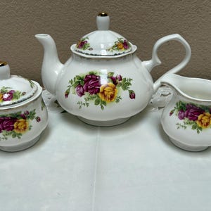 May include: A white porcelain tea set consisting of a teapot with a lid, a sugar bowl with a lid, and a creamer. Each piece features a floral pattern of deep magenta and yellow roses with green leaves and gold trim along the edges and handles.