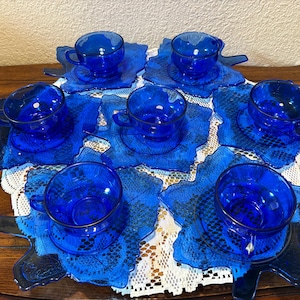 May include: A set of six cobalt blue glass teacups and saucers. The cups have handles and sit on leaf-shaped saucers. The set is arranged on a white lace doily and a dark wooden table. The teacups and saucers are made of glass.