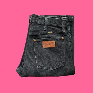 Puede incluir: Vaqueros Wrangler negros con un parche de cuero marr&oacute;n en el bolsillo trasero. Los vaqueros tienen un dise&ntilde;o cl&aacute;sico de cinco bolsillos, trabillas para el cintur&oacute;n y cierre de bot&oacute;n y cremallera. El logo de Wrangler es visible.