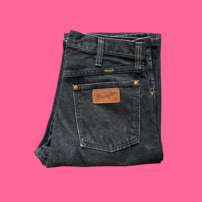 Puede incluir: Vaqueros Wrangler negros con un parche de cuero marr&oacute;n en el bolsillo trasero. Los vaqueros tienen un dise&ntilde;o cl&aacute;sico de cinco bolsillos, trabillas para el cintur&oacute;n y cierre de bot&oacute;n y cremallera. El logo de Wrangler es visible.