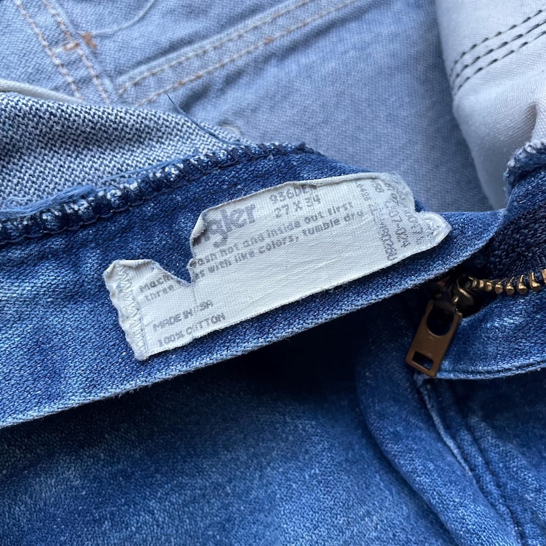 May include: Close-up of blue denim jeans with a zipper and a tag. The tag reads "Wrangler" and includes washing instructions and size 27 x 34. The jeans are made of 100% cotton and are made in the USA.