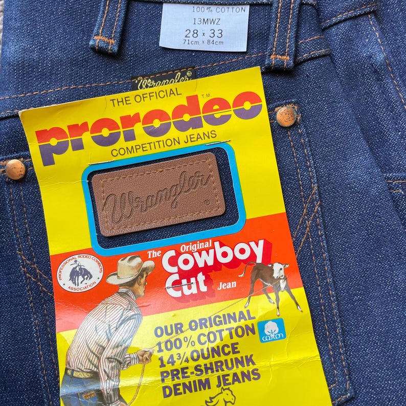 Op de afbeelding: Close-up van een donkerblauwe Wrangler ProRodeo Competition Jeans. De jeans heeft een label met de tekst "100% COTTON, 13MWZ, 28x33" en een geel label met "Cowboy Cut Jean" en "14 3/4 ounce".