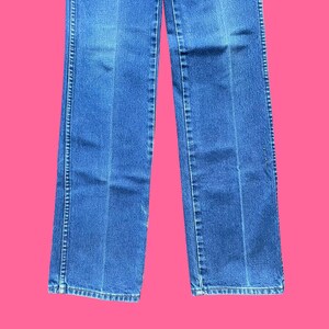 May include: Blue denim jeans with a straight leg cut. The jeans have a classic five-pocket design, a zipper fly, and a button closure. The jeans are a medium wash and have a slight fade.