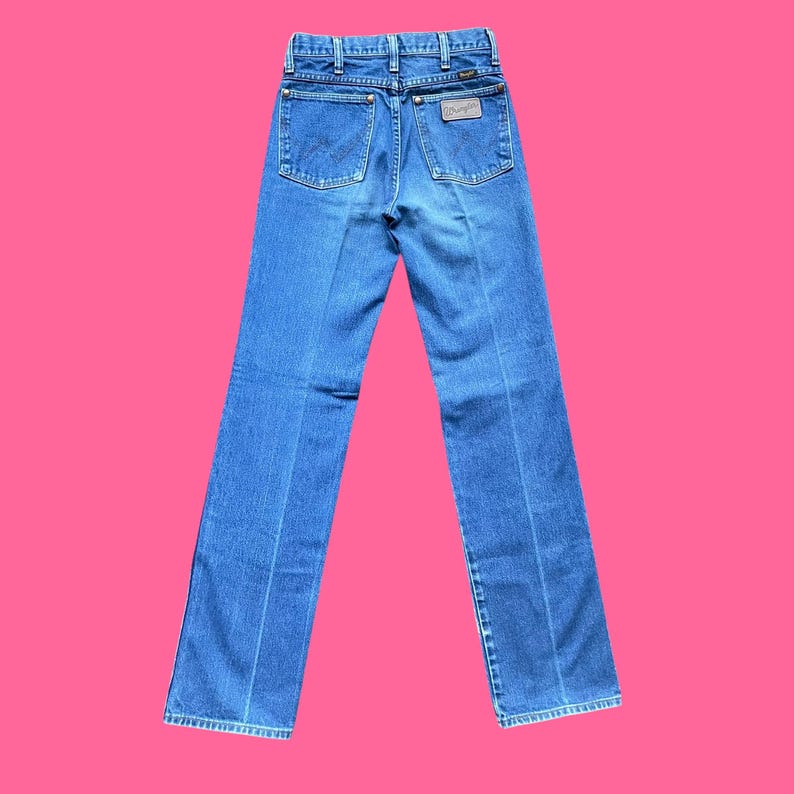 May include: Blue denim jeans with a classic five-pocket design. The jeans feature a straight leg cut and a brown leather patch with the brand name on the back. The jeans are displayed against a bright pink background.