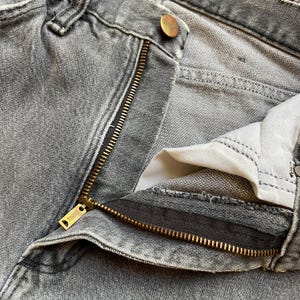 May include: Close-up of gray denim jeans with a partially unzipped brass zipper. The jeans have a button closure and a small brass button. The zipper pull has the word "THINGS" engraved on it. The jeans are faded and worn.