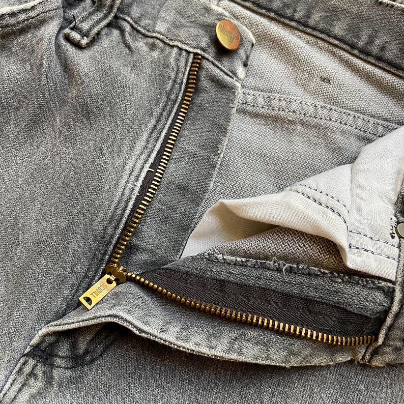 May include: Close-up of gray denim jeans with a partially unzipped brass zipper. The jeans have a button closure and a small brass button. The zipper pull has the word "THINGS" engraved on it. The jeans are faded and worn.