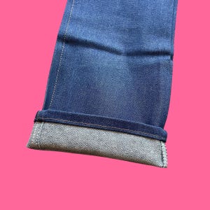 Op de afbeelding: Close-up van een blauwe denim jeans met een omgeslagen zoom. De jeans heeft een klassiek vijf-zakken ontwerp en is gemaakt van een duurzame denim stof. De zoom is omgeslagen en onthult de lichtere binnenkant van de denim.
