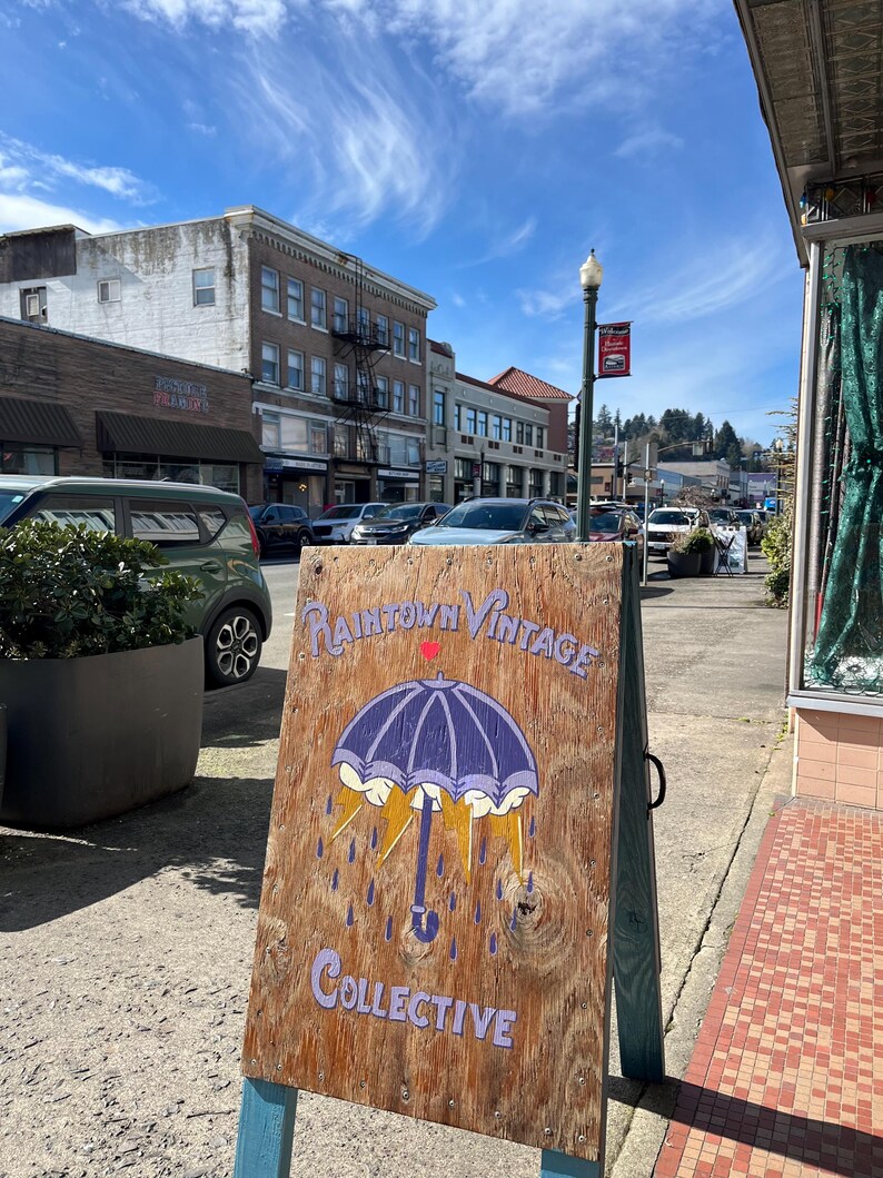 May include: Wooden sandwich board sign with the text "Raintown Vintage Collective" and an umbrella illustration. The sign is on a sidewalk in front of a building with other buildings and cars in the background. The sky is blue with clouds.