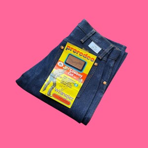 Op de afbeelding: Donkerblauwe Wrangler Cowboy Cut jeans met een geel label. Het label leest "prorodeo COMPETITION JEANS" en "Cowboy Cut". De jeans is maat 28-33 en gemaakt van 100% katoen, 14-ounce voorgekrompen denim. Het label zegt ook "FULL 1 YEAR WARRANTY."