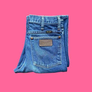 May include: Folded blue denim jeans with the Wrangler logo on the back pocket. The jeans have a classic five-pocket design, belt loops, and copper-colored rivets. The jeans are folded neatly against a pink background.