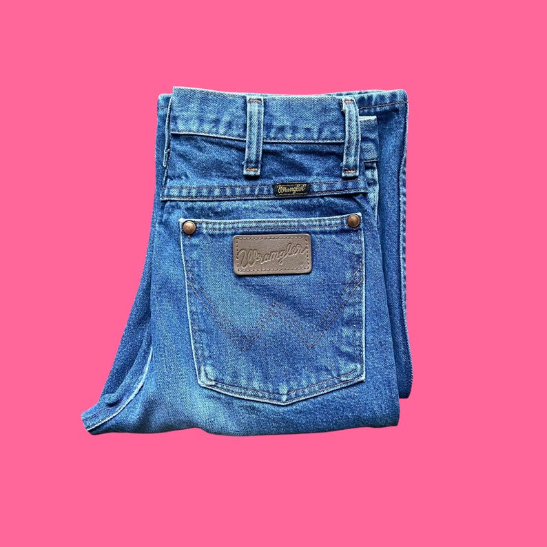 May include: Folded blue denim jeans with the Wrangler logo on the back pocket. The jeans have a classic five-pocket design, belt loops, and copper-colored rivets. The jeans are folded neatly against a pink background.