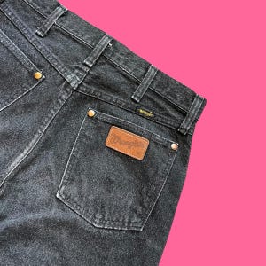 Puede incluir: Primer plano de la parte trasera de unos vaqueros Wrangler negros. Los vaqueros tienen un dise&ntilde;o cl&aacute;sico de cinco bolsillos con un parche de cuero marr&oacute;n con el logotipo de Wrangler. Los vaqueros tienen remaches color lat&oacute;n y una cintura negra.