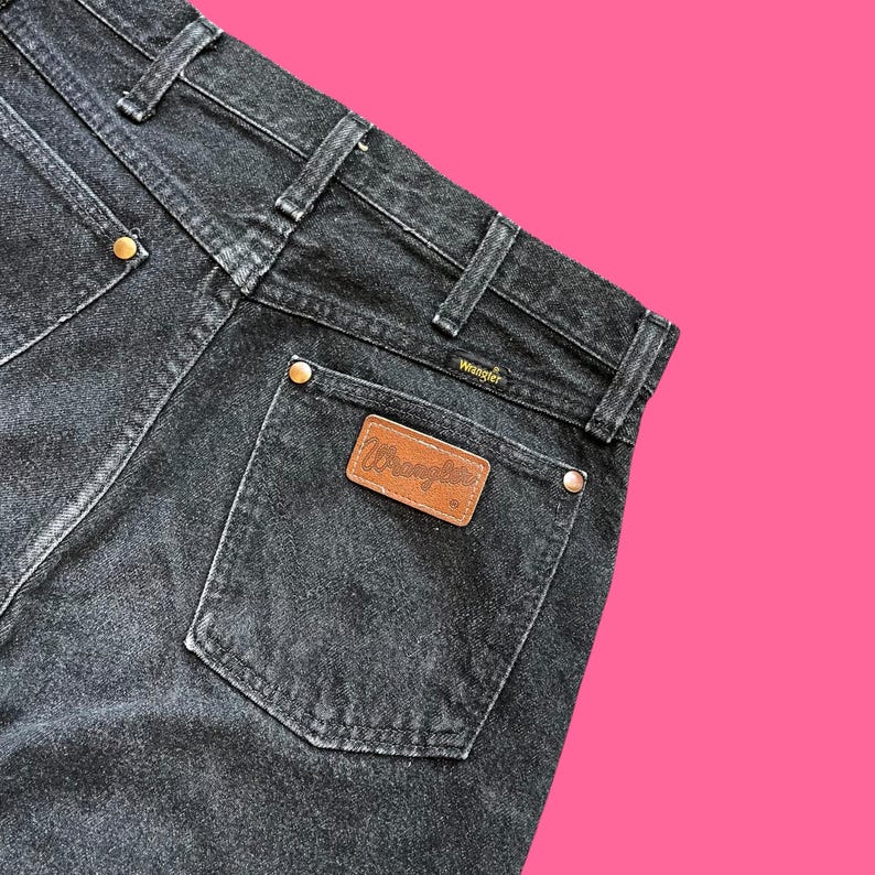 Puede incluir: Primer plano de la parte trasera de unos vaqueros Wrangler negros. Los vaqueros tienen un dise&ntilde;o cl&aacute;sico de cinco bolsillos con un parche de cuero marr&oacute;n con el logotipo de Wrangler. Los vaqueros tienen remaches color lat&oacute;n y una cintura negra.