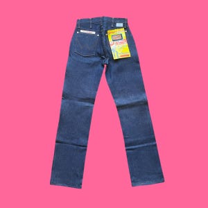 Op de afbeelding: Donkerblauwe denim jeans met een rechte pijp. De jeans hebben een klassiek ontwerp met vijf zakken en een Wrangler-label op de achterzak. Een geel en wit label is aan de achterkant bevestigd. De jeans worden gepresenteerd tegen een roze achtergrond.