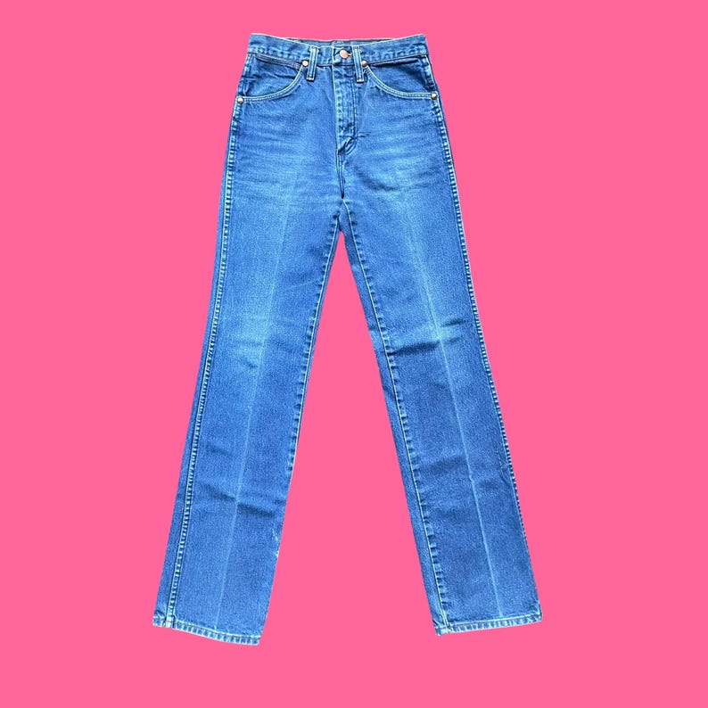 May include: Blue denim jeans with a classic five-pocket design. The jeans have a high waist and a straight leg fit. The denim is a medium wash with subtle fading and a slight sheen. The jeans are displayed against a pink background.