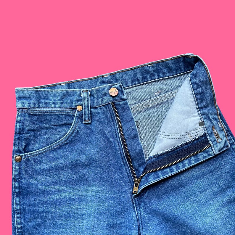 May include: Close-up of blue denim jeans with a partially unzipped fly. The jeans have a classic five-pocket design, with a button closure and metal rivets. The denim fabric is a medium wash, showing some wear.