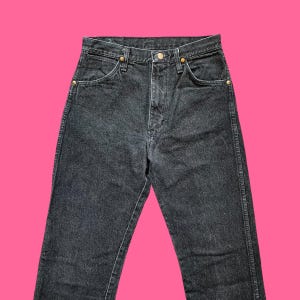 Puede incluir: Vaqueros de mezclilla negros con bragueta de botones y dise&ntilde;o cl&aacute;sico de cinco bolsillos. Los vaqueros tienen remaches dorados y corte recto. La prenda se muestra sobre un fondo rosa brillante.