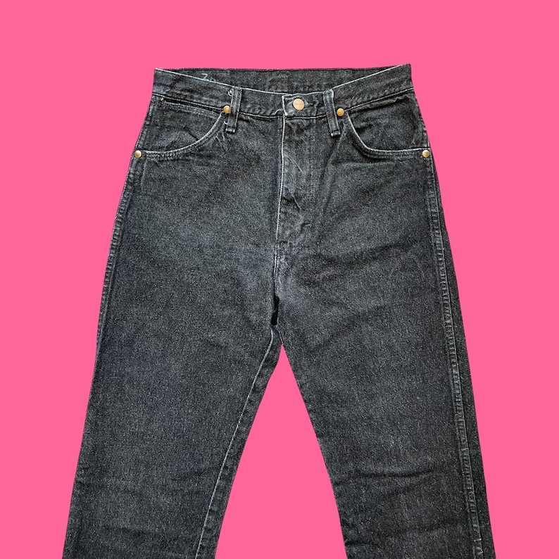 Puede incluir: Vaqueros de mezclilla negros con bragueta de botones y dise&ntilde;o cl&aacute;sico de cinco bolsillos. Los vaqueros tienen remaches dorados y corte recto. La prenda se muestra sobre un fondo rosa brillante.