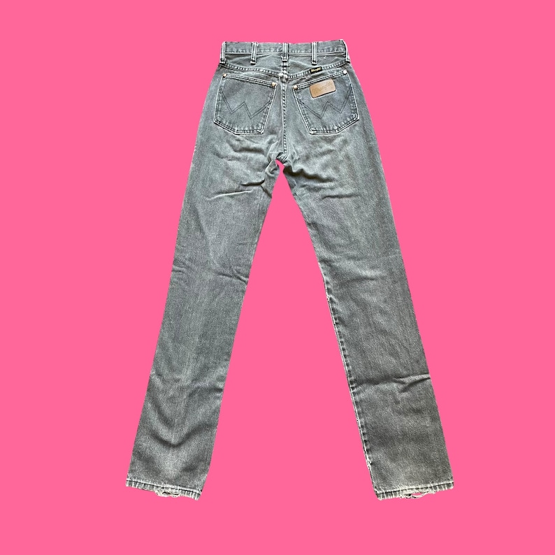 May include: Gray denim jeans with a classic five-pocket design. The jeans feature a straight leg cut and a brown leather patch on the back. The Wrangler logo is visible on the patch and back pockets.