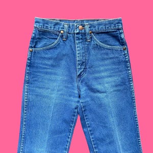 May include: Close-up of blue denim jeans. The jeans have a classic five-pocket design with a button and zipper closure. The denim is a medium wash with subtle fading and wear. The jeans are high-waisted and have a straight leg.