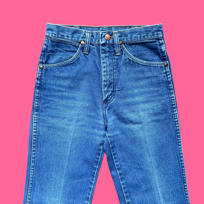 May include: Close-up of blue denim jeans. The jeans have a classic five-pocket design with a button and zipper closure. The denim is a medium wash with subtle fading and wear. The jeans are high-waisted and have a straight leg.
