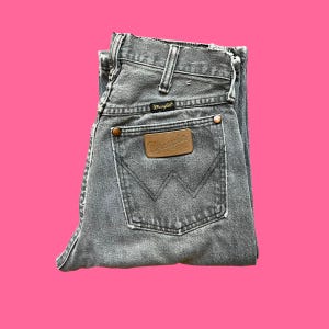 May include: Folded gray Wrangler jeans with a brown leather patch and copper rivets. The jeans have a classic five-pocket design and a faded, worn look. The Wrangler logo is visible on the patch and the back pocket.