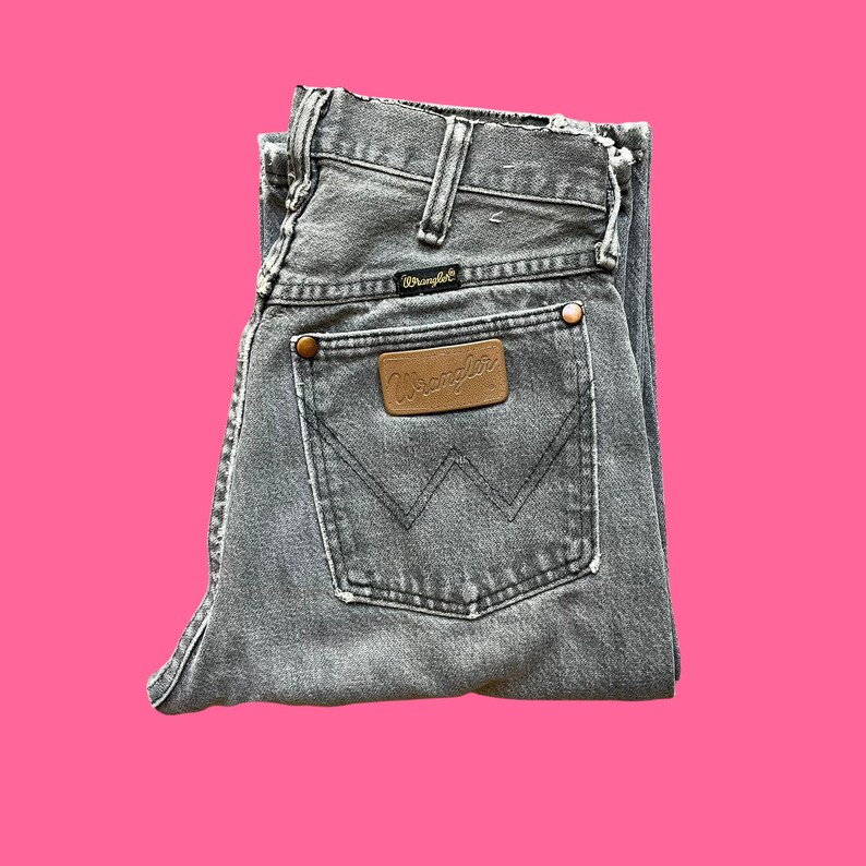 May include: Folded gray Wrangler jeans with a brown leather patch and copper rivets. The jeans have a classic five-pocket design and a faded, worn look. The Wrangler logo is visible on the patch and the back pocket.