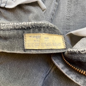 May include: Close-up of gray Wrangler jeans with a zipper and a tag. The tag reads "Wrangler" and includes size and length information. The jeans are made in the USA.