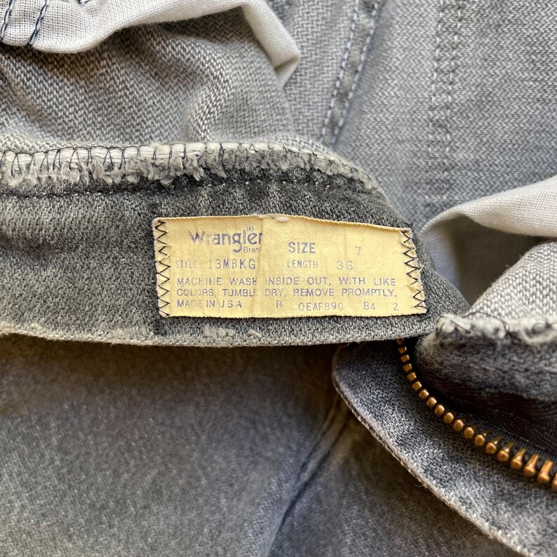 May include: Close-up of gray Wrangler jeans with a zipper and a tag. The tag reads "Wrangler" and includes size and length information. The jeans are made in the USA.
