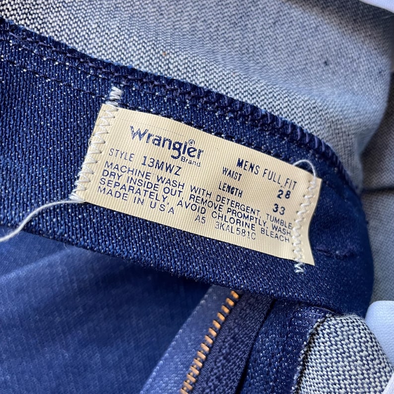Op de afbeelding: Close-up van een Wrangler-merklabel op donkerblauwe denim jeans. Het label geeft een taillemaat van 71 cm en een lengte van 84 cm aan. Het label bevat ook wasvoorschriften en de tekst "MADE IN USA".