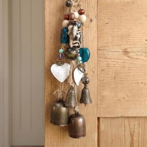 May include: A handcrafted wind chime with a rustic design. It features a jute rope strung with beads in various colours, including white, brown, and teal. Below, heart-shaped pendants and small bronze bells hang, creating a charming, handcrafted look.