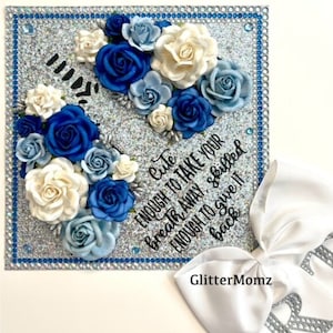 Könnte beinhalten: Eine Abschluss-Kappe, die mit blauen und weißen Blumen und Strasssteinen verziert ist. Die Kappe hat einen silbernen Glitzer-Hintergrund und den Text "Cute enough to take your breath away, skilled enough to give it back" ist darauf geschrieben.