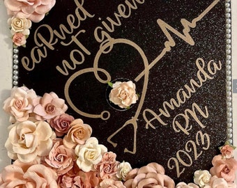 May include: A black graduation cap decorated with the words "earned not given", a stethoscope heart design, the name "Amanda RN 2023", and an assortment of pink and cream-colored paper flowers. The cap is trimmed with pearls.