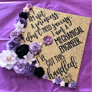 May include: Gold graduation cap decorated with purple, white, and black flowers. The cap has a gold glitter background and black lettering that reads "I am not a princess. I don't need saving. I am a Mechanical Engineer. I've got this handled. BSME 2021"