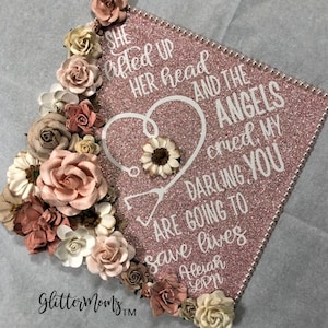 May include: A pink glitter graduation cap decorated with white text and a stethoscope inside a heart. The text reads "She lifted up her head and the angels cried, my darling, you are going to save lives". The cap is also decorated with pink, white, and brown paper flowers.